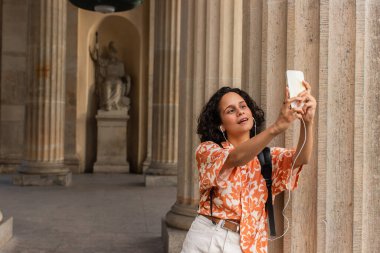 curly young woman in wired earphones taking selfie with statue while holding smartphone 