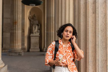 curly young woman adjusting wired earphones while holding smartphone near blurred statue in berlin 
