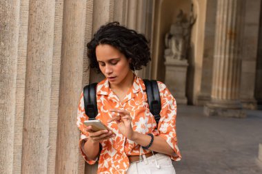curly young woman in wired earphones using smartphone near blurred statue in berlin 