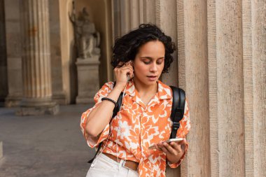 young woman adjusting wired earphones while using smartphone near blurred statue in berlin 