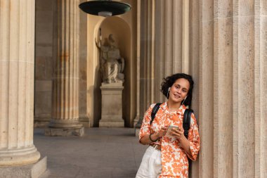 happy young woman in wired earphones listening music and holding smartphone near statue in berlin 