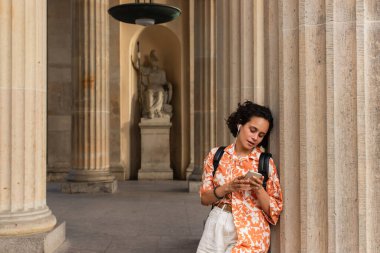 pretty young woman in wired earphones listening music and holding smartphone near statue in berlin 