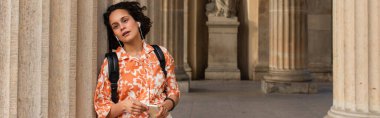 traveler in wired earphones listening music and holding smartphone near statue in berlin, banner