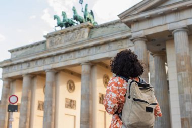 back view of curly tourist with backpack standing near brandenburg gate in berlin 