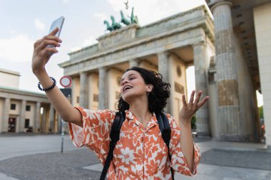 curly tourist with backpack taking selfie on smartphone near brandenburg gate in berlin 
