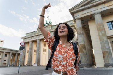 curly tourist with backpack waving hand near brandenburg gate in berlin 
