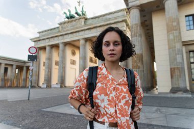 curly young woman with backpack standing near brandenburg gate in berlin 