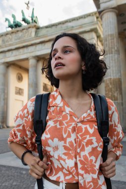 curly tourist with backpack looking away near brandenburg gate in berlin 