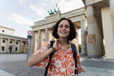 cheerful young woman with backpack standing near brandenburg gate in berlin 
