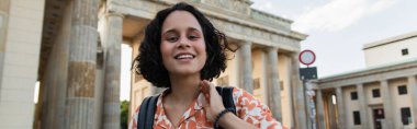 cheerful young woman with backpack standing near brandenburg gate in berlin, banner