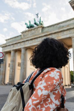 back view of curly traveler with backpack standing near brandenburg gate in berlin  clipart