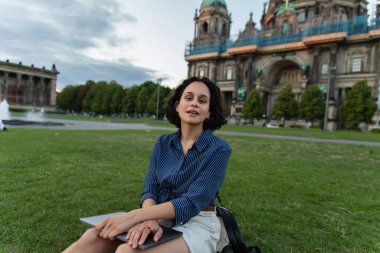 curly young woman sitting on grass with laptop near blurred cathedral in berlin 