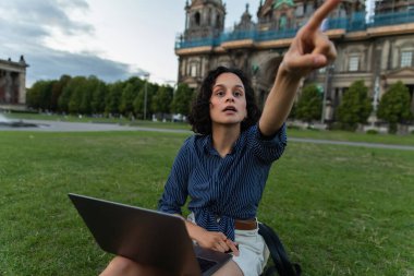 curly young woman with laptop pointing with finger and sitting on grass near blurred cathedral in berlin 