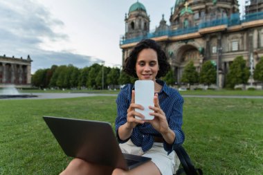 smiling woman sitting with laptop and taking selfie on smartphone near cathedral in berlin