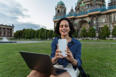 happy woman sitting with laptop and taking selfie on smartphone near cathedral in berlin
