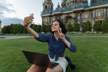 cheerful freelancer sitting with laptop and taking selfie on smartphone and showing peace sign near cathedral in berlin