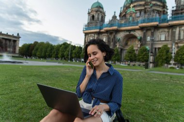young woman sitting with laptop and talking on smartphone near cathedral in berlin