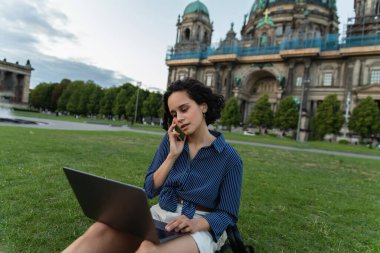 curly young woman sitting with laptop and talking on smartphone near cathedral in berlin