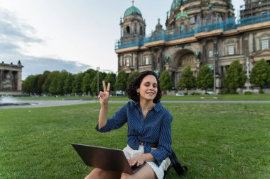 happy young woman with laptop showing peace sign and sitting on grass near blurred cathedral in berlin 