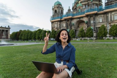 amazed young woman with laptop showing peace sign and sitting on grass near blurred cathedral in berlin 