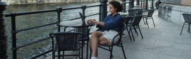 young freelancer using laptop while sitting in cafe terrace near river in berlin, banner