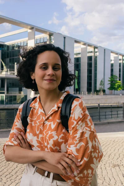 smiling young woman with backpack standing near modern contemporary building in berlin