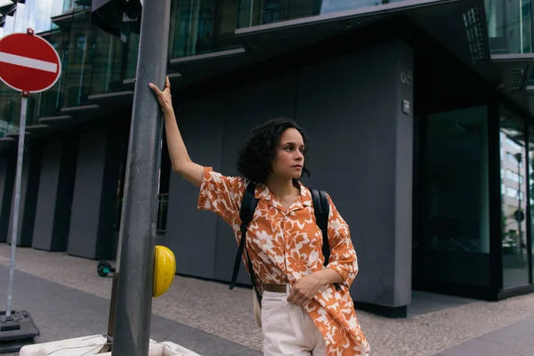 curly young woman with backpack standing on urban street in berlin 