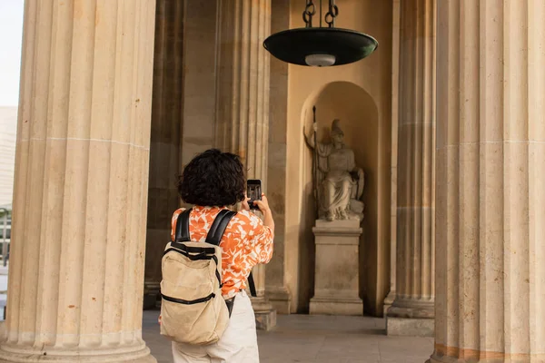 back view of curly woman with backpack taking photo of statue on smartphone 