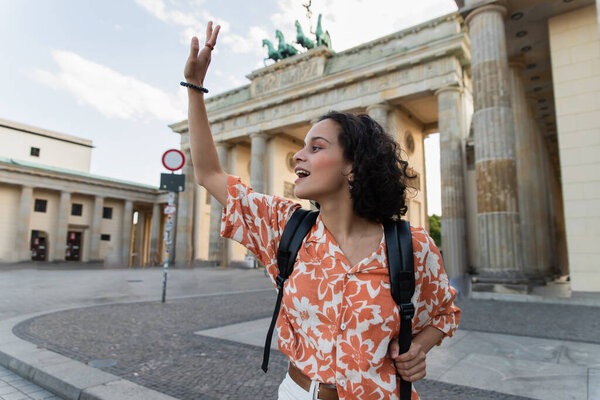 excited tourist with backpack waving hand near brandenburg gate in berlin 
