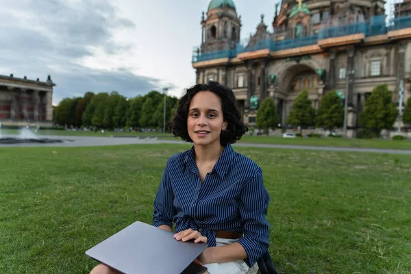 happy young woman holding laptop and sitting on grass near blurred cathedral in berlin 
