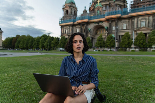 shocked young woman holding laptop and sitting on grass near blurred cathedral in berlin 