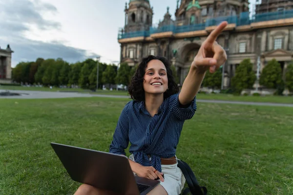 cheerful young woman with laptop pointing with finger and sitting on grass near blurred cathedral in berlin
