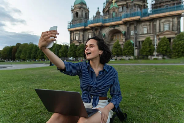 amazed woman sitting with laptop and taking selfie on smartphone near cathedral in berlin