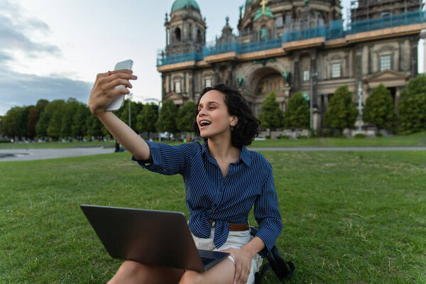 amazed woman sitting with laptop and taking selfie on smartphone near cathedral in berlin