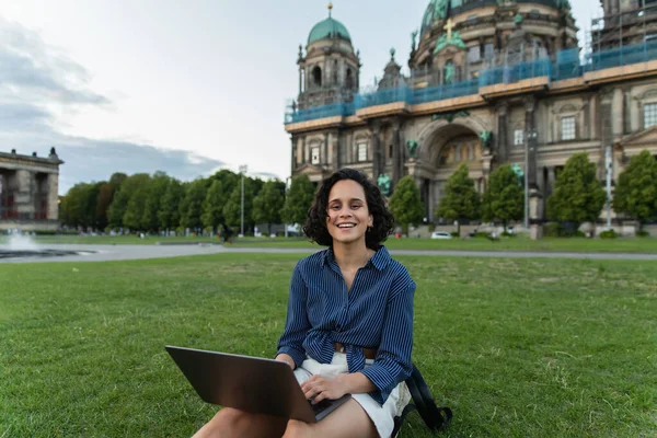 cheerful young woman holding laptop and sitting on grass near blurred cathedral in berlin 