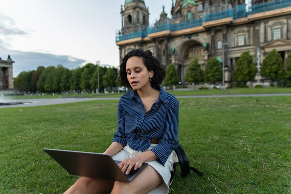 young woman using laptop and sitting on lawn near blurred cathedral in berlin
