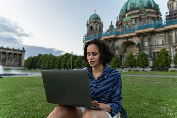 emotional young woman using laptop and sitting on lawn near blurred cathedral in berlin