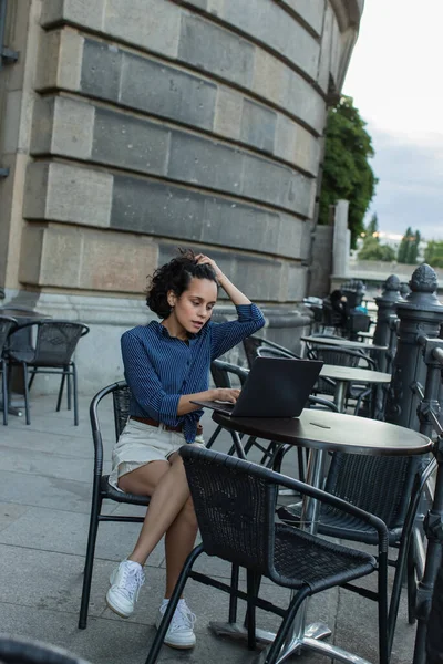 young freelancer adjusting curly hair and using laptop while sitting on summer terrace in berlin 