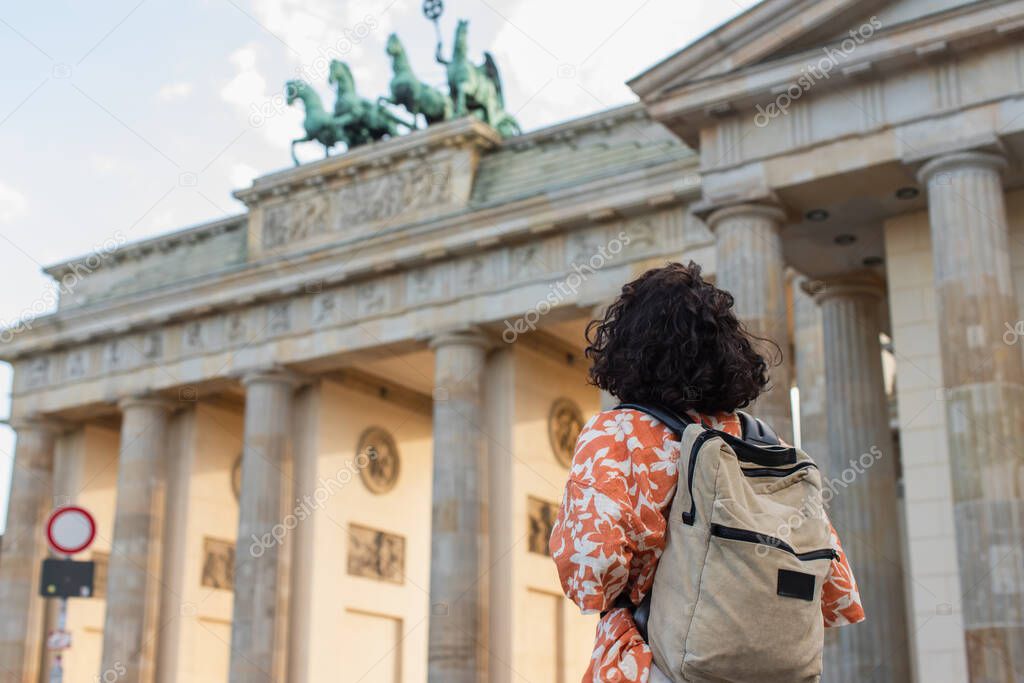 back view of curly tourist with backpack standing near brandenburg gate in berlin 