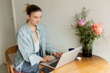 Genç bir kadın evinde çiçek ve fincanın yanında laptop kullanıyor. 