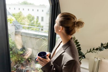 Evin balkon kapısının yanında ceketli genç bir kadın bardak tutuyor. 