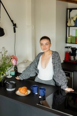 Woman in knitted cardigan standing near croissant and cups in kitchen  clipart