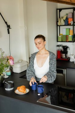 Elinde çay poşeti olan hırkalı bir kadın mutfakta ayçöreği tutuyor. 