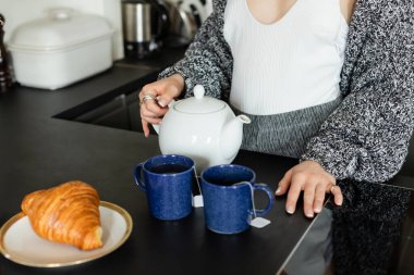 Örgü hırka giymiş bir kadının elinde çaydanlık ve evde bulanık kruvasan tutarken görüntüsü. 