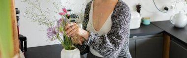 Cropped view of woman in cardigan putting bouquet in vase at home, banner  clipart