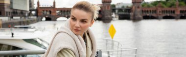 Young woman in trench coat standing on pier with Oberbaum bridge at background in Germany, banner  clipart