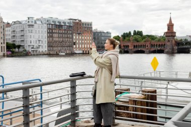Side view of young woman taking photo on pier in Berlin  clipart