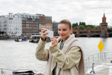 Trençkotlu gülümseyen kadın Berlin 'de cep telefonuyla fotoğraf çekiyor. 