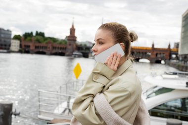 Berlin 'de nehir kenarında trençkotlu bir kadının cep telefonuyla konuşmasının yan görüntüsü. 