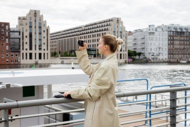 Trençkotlu bir kadının Berlin 'de nehir kenarında cep telefonuyla fotoğraf çekerken yan görüntüsü. 